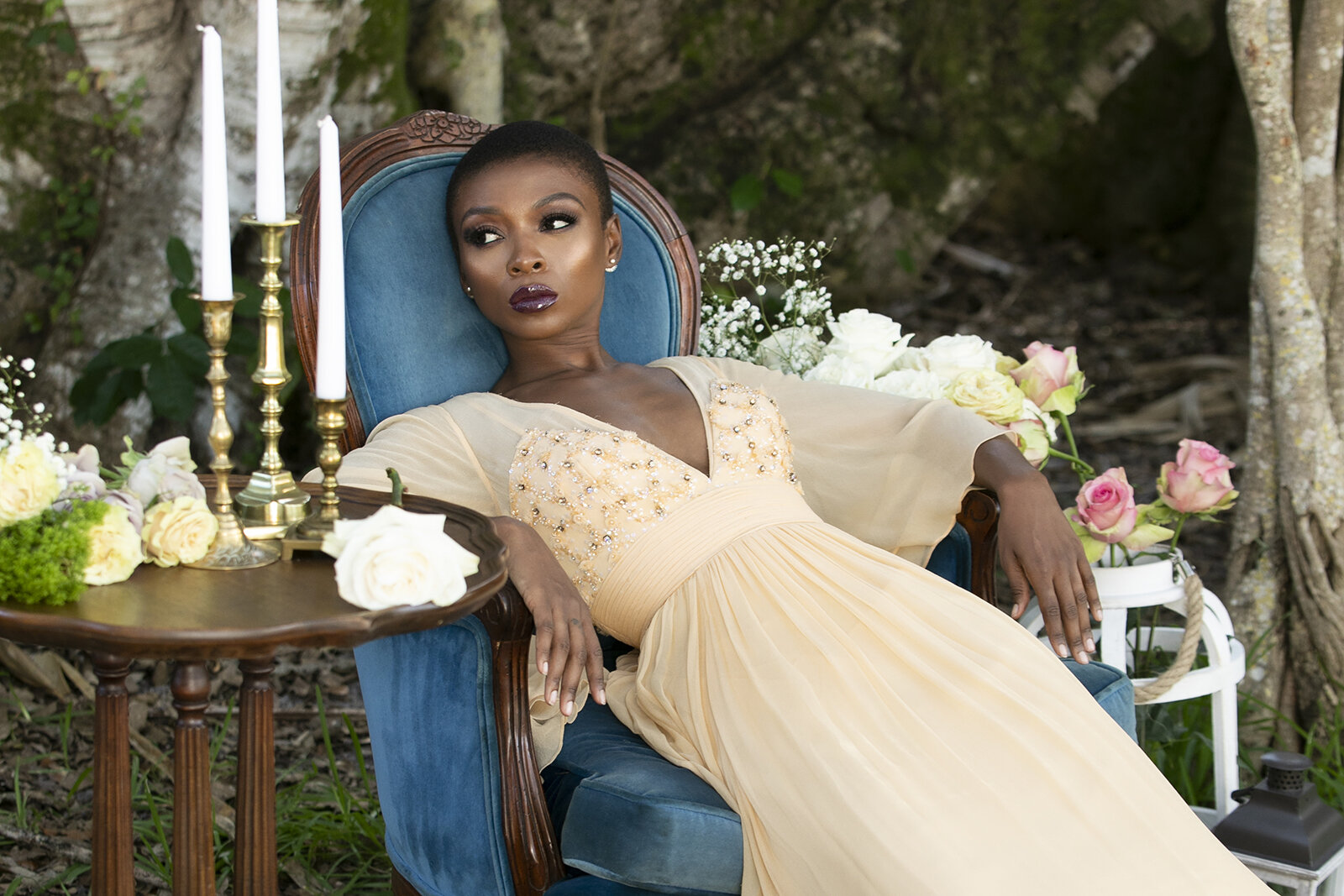 Model in a pale gown reclining in a blue chair among flowers and candles