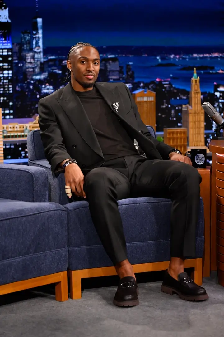 Guest in a black suit seated on a blue armchair on a late-night talk show set